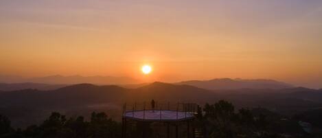 A sunset view from a circular observation deck with a person standing, overlooking a range of mountains.