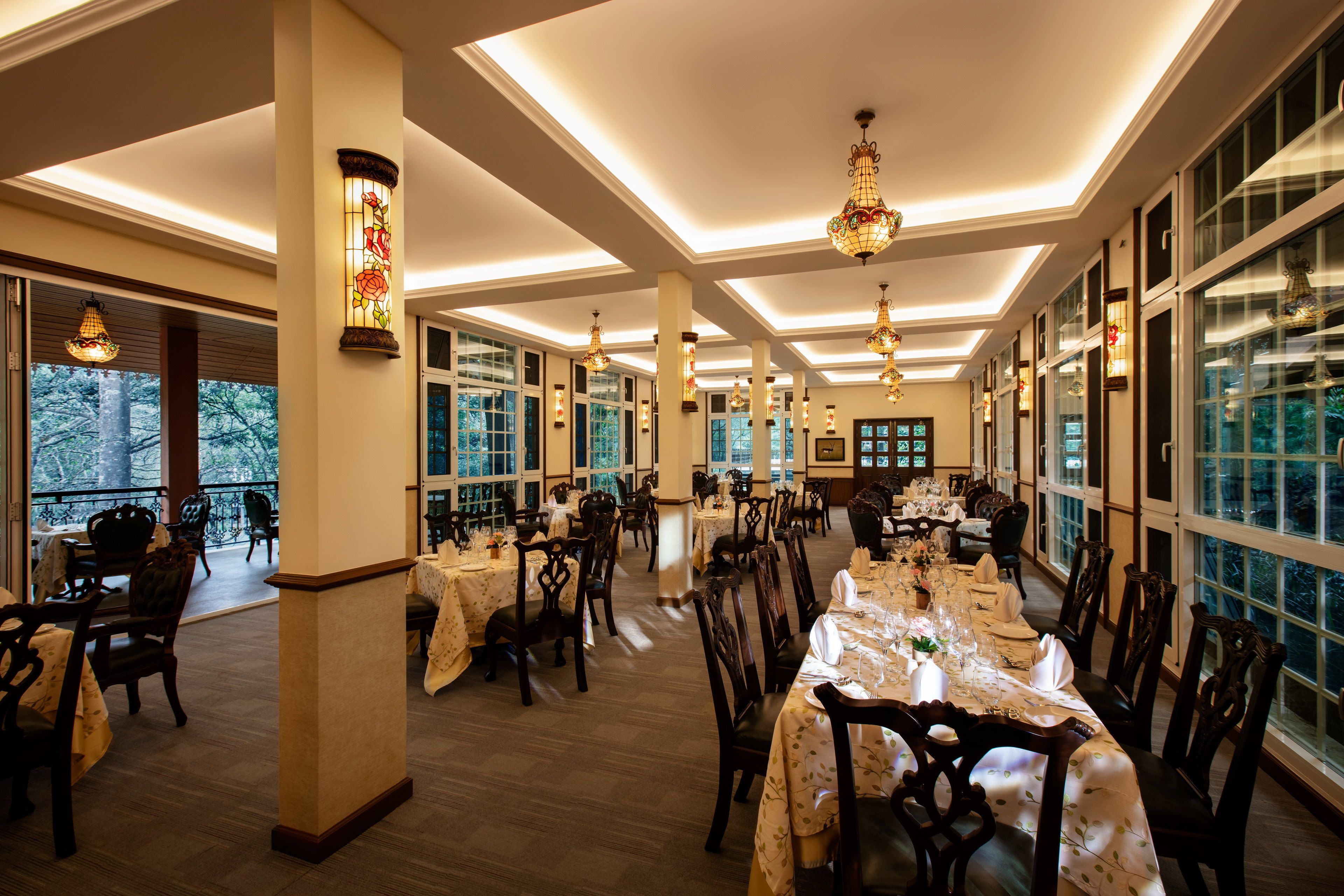 Un comedor amplio con varias mesas dispuestas para comer, ventanas grandes y lámparas decorativas.