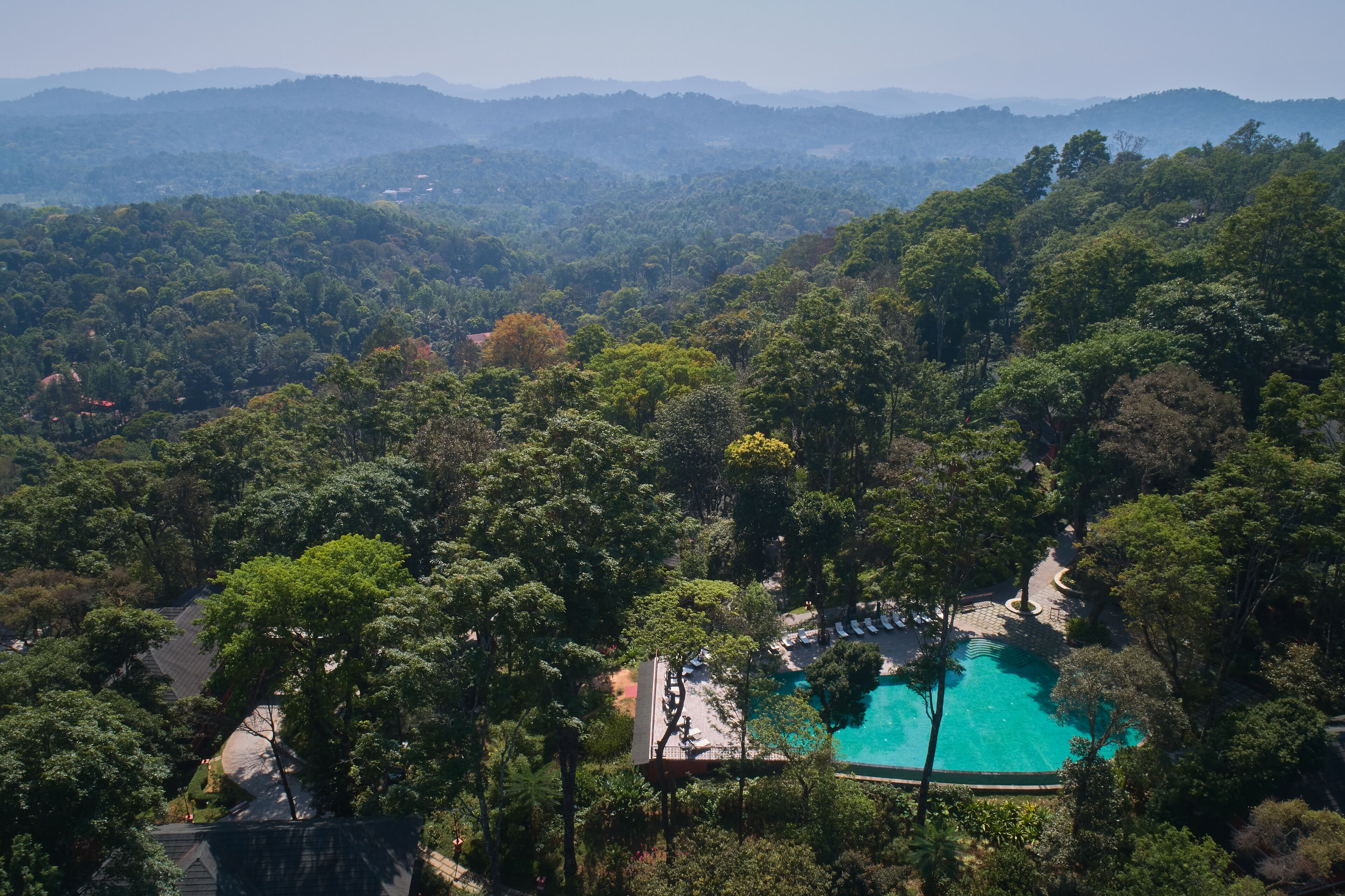 An aerial view of a resort with a swimming pool surrounded by dense forest and hills in the background.