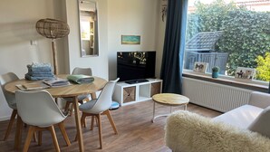 A modern living room with a dining area, a TV, and a view of greenery outside. - Holiday summer style house, family or couple. With privacy/little garden/ sea (Noordwijk aan Zee)