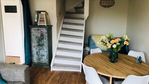 A dining area with a wooden table and white chairs, a staircase leading to another level, and a vintage cabinet with a picture frame. - Holiday summer style house, family or couple. With privacy/little garden/ sea (Noordwijk aan Zee)