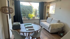 A cozy living room with a round wooden table, two white chairs, a white sofa with a fluffy throw, and a window with curtains. - Holiday summer style house, family or couple. With privacy/little garden/ sea (Noordwijk aan Zee)