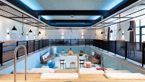 Una piscina interior moderna con techo azul, lámparas colgantes negras y varios asientos y equipos de ejercicio.