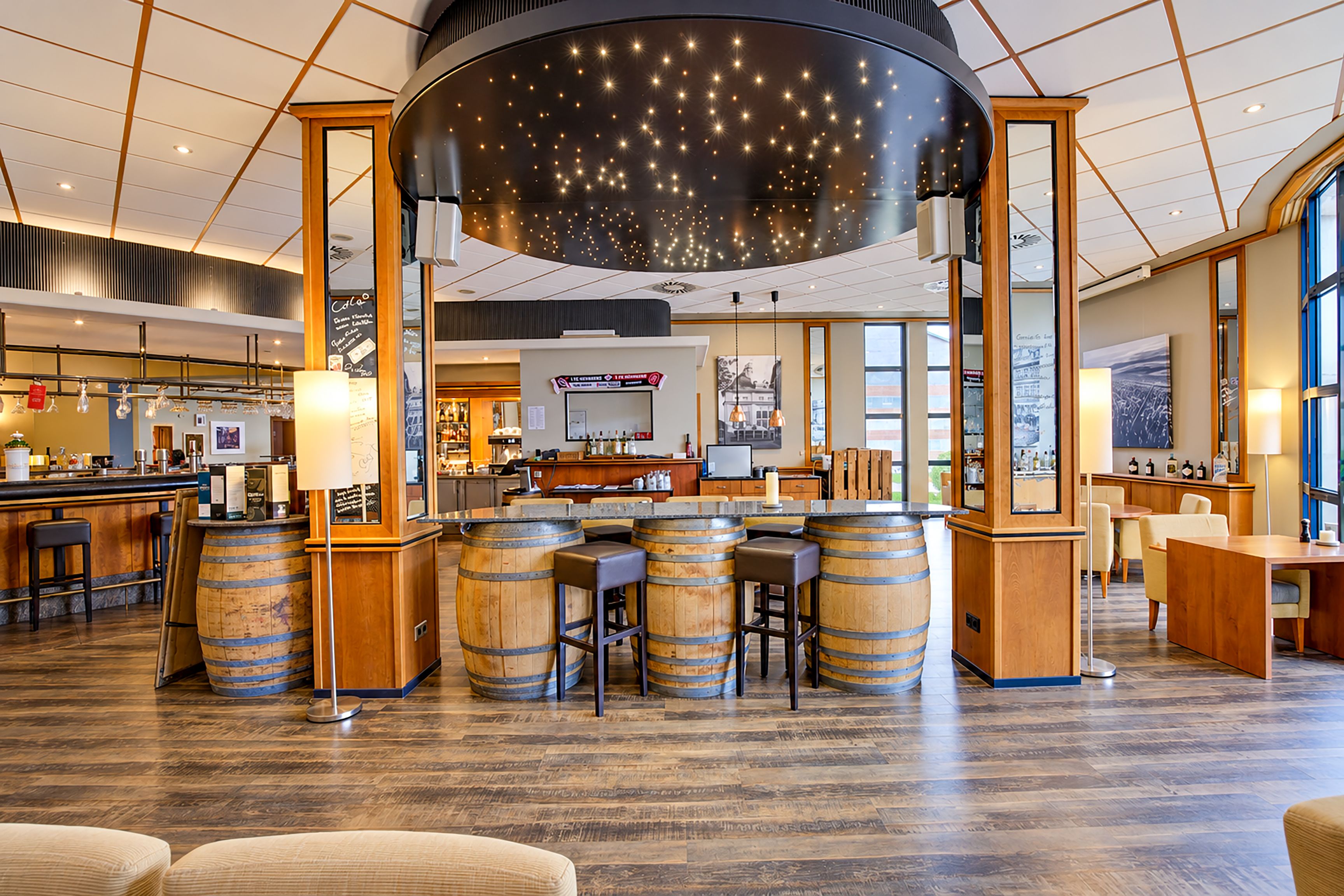 A bar area with wooden barrels, a large hanging light fixture, and a counter with stools.