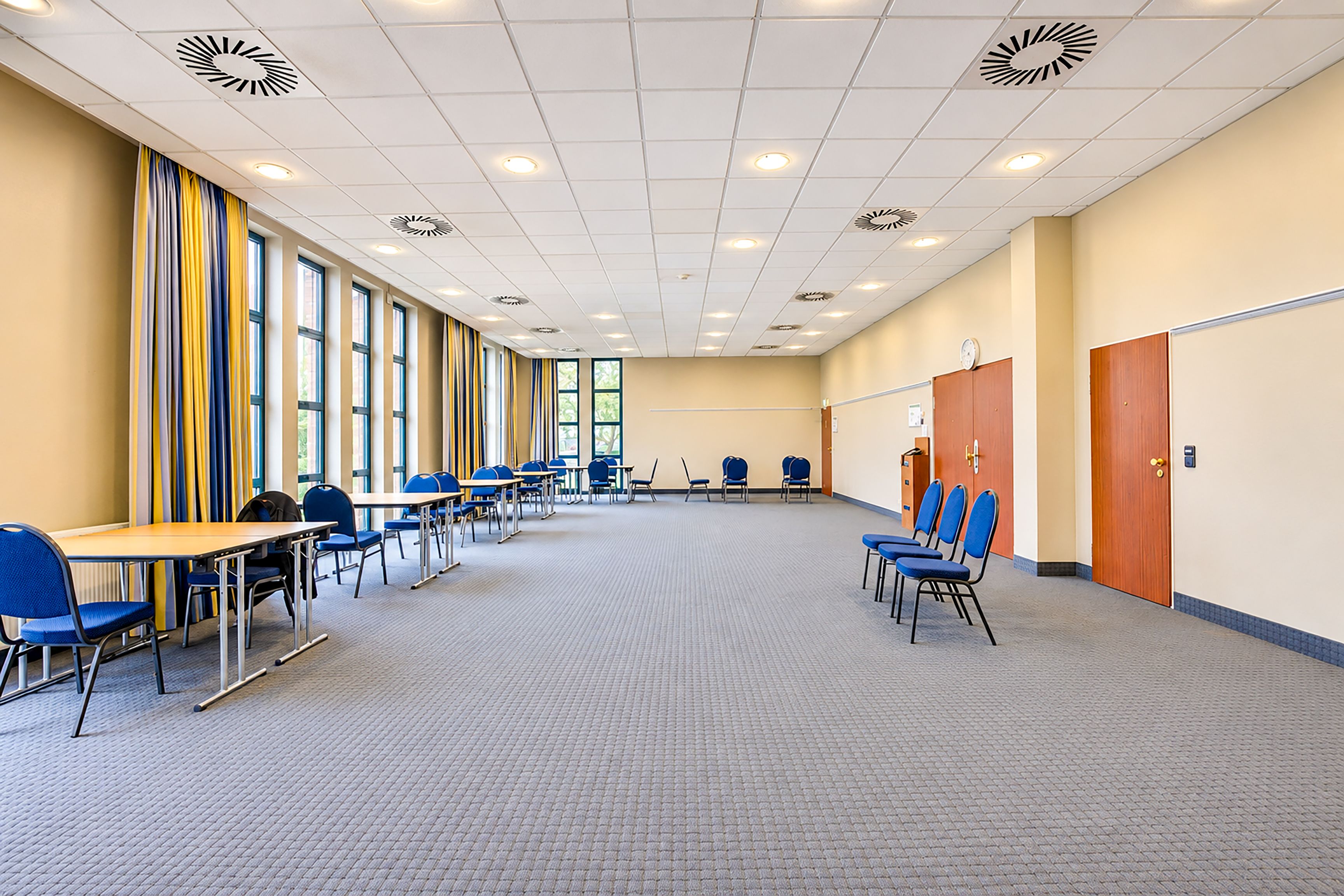 A conference room with rows of tables and chairs, large windows, and a tiled ceiling.