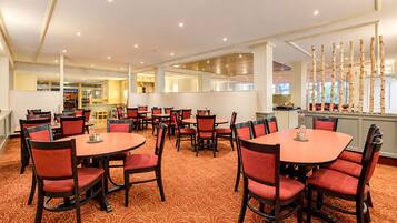 Um espaço de refeições de hotel com mesas e cadeiras redondas, um carpete estampado e vista para a cozinha através de um painel de vidro.