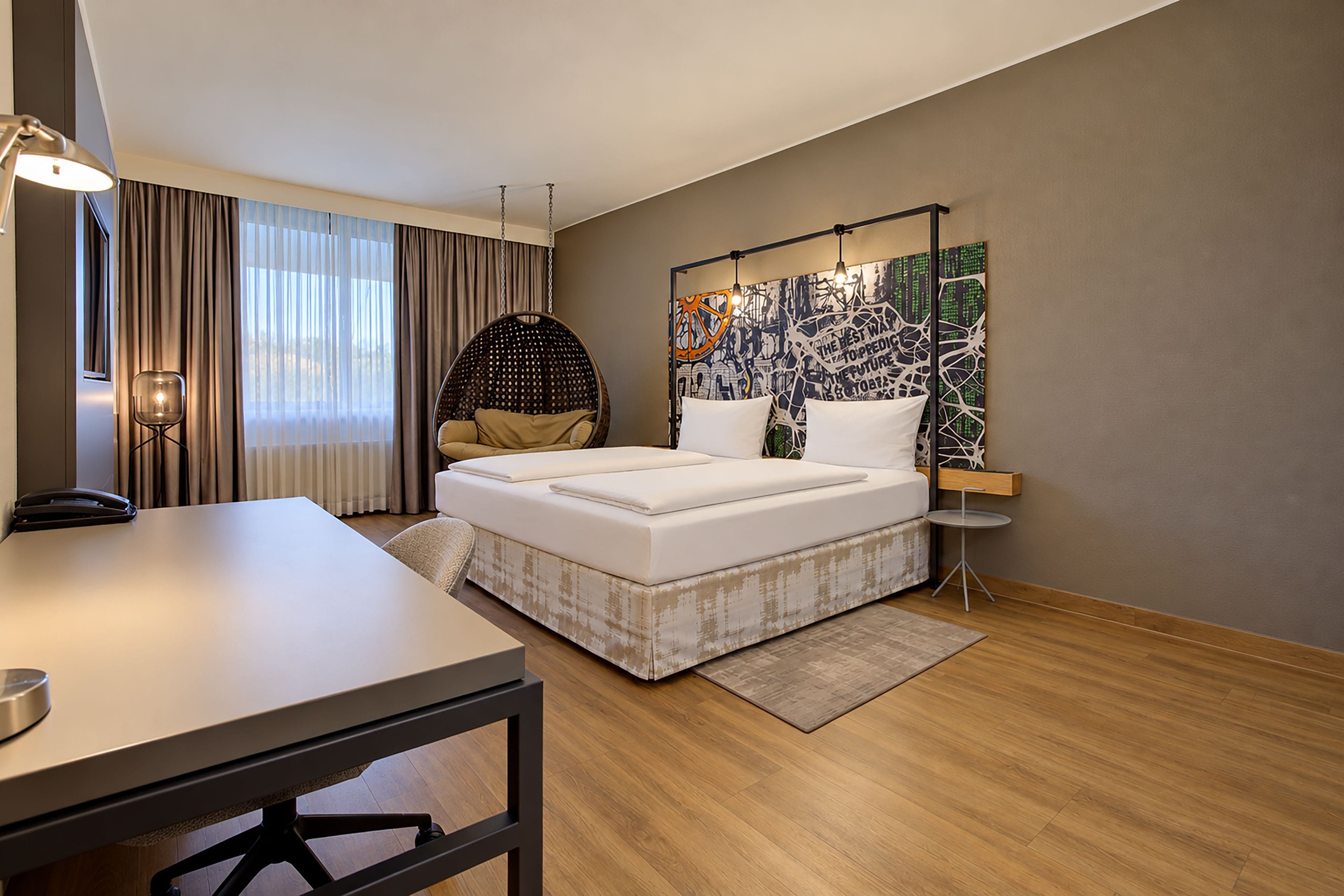 A modern hotel room with a bed, desk, chair, and artwork on the wall.