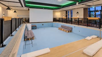 Une piscine intérieure moderne avec un sol carrelé de bleu, une terrasse en bois et des espaces aménagés pour s’asseoir.