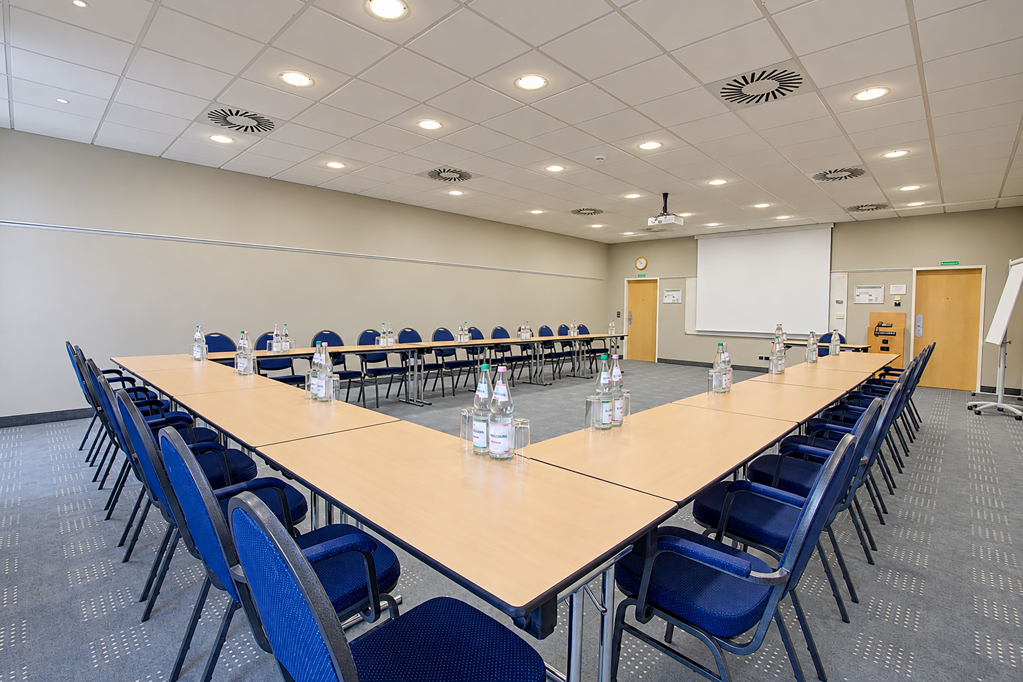 Un salón de conferencias con una larga mesa, sillas azules, botellas de agua, una pantalla de proyector y una pizarra.