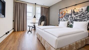 Hotelkamer met een groot bed, een bureau en een stoel. Er is een raam met gordijnen, een hanglamp en een muur met een stadsgezichtsmural.