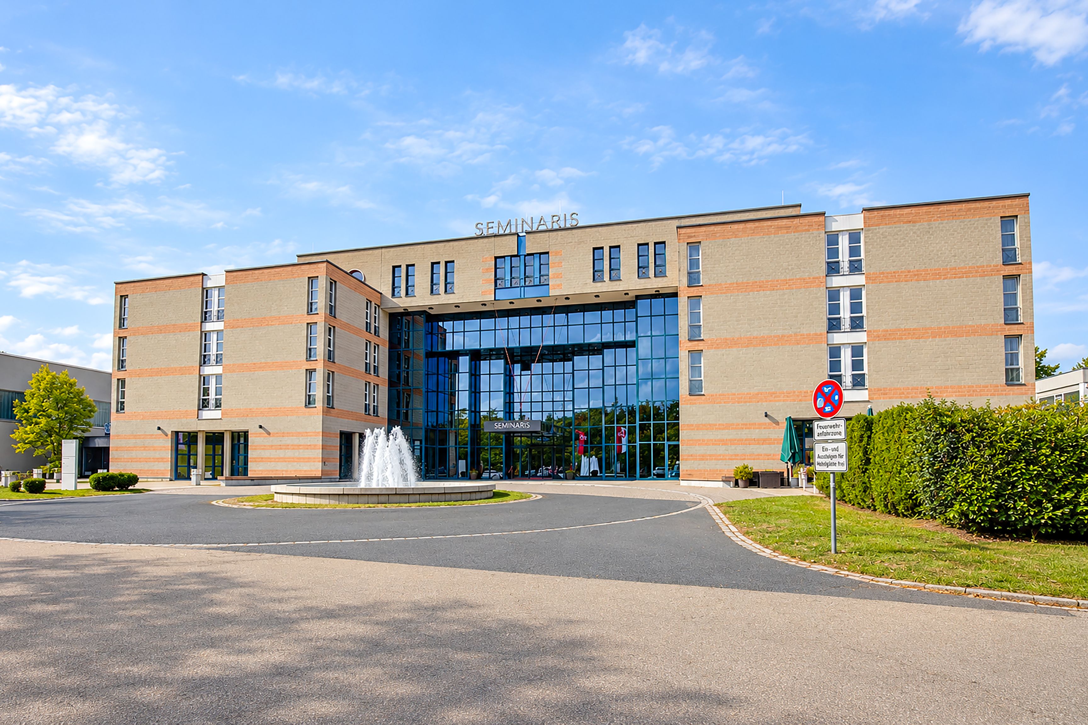 Ein modernes Gebäude mit einem markanten Eingangsbereich und einer Springbrunnenanlage vor der Fassade, bewölkter Himmel.