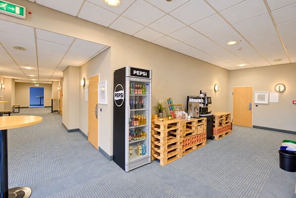 A room with a vending machine, wooden crates, a coffee machine, and a blue door. - Seminaris Sunday Hotel Nürnberg (Nuremberg)