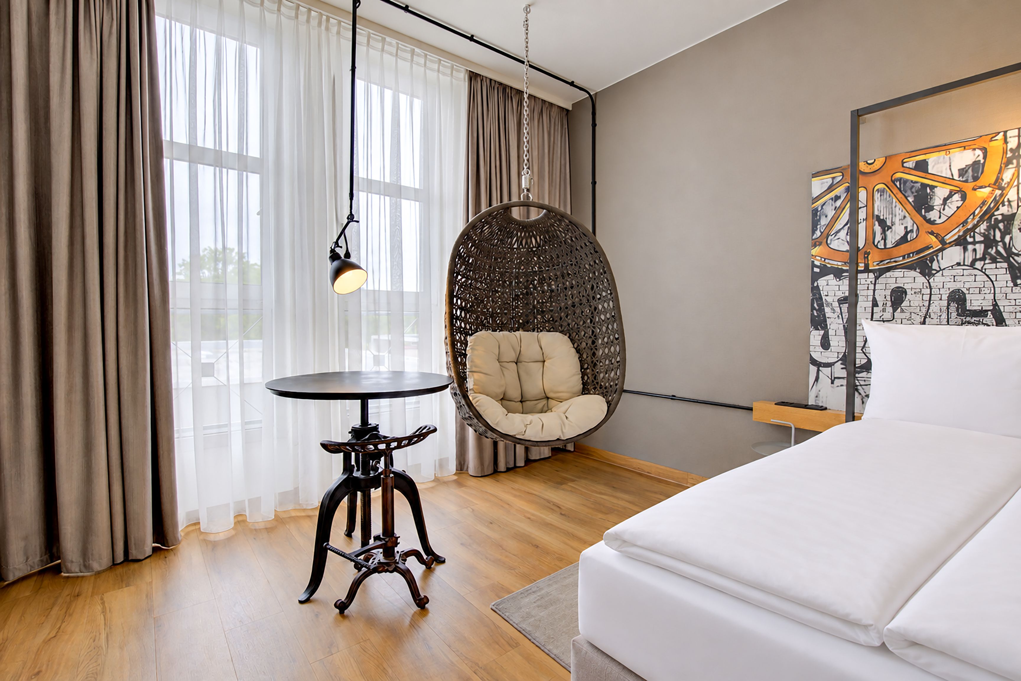 Una camera d'albergo moderna con un letto, una poltrona rotonda in vimini, un tavolino e un quadro appeso al muro.
