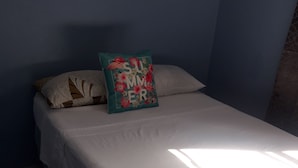 Una cama con colchón blanco y almohada estampada, suelo de baldosas y pared azul.