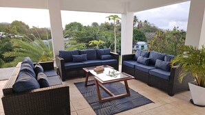 Un patio al aire libre con mobiliario de mimbre, una mesa de centro de vidrio y vistas al entorno verde.