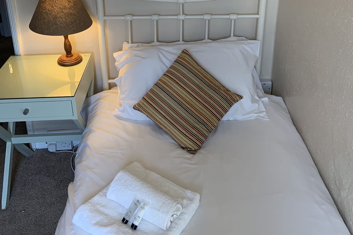 A neatly made bed with white linens, a striped pillow, and a folded towel. A bedside lamp is on the nightstand beside the bed.
