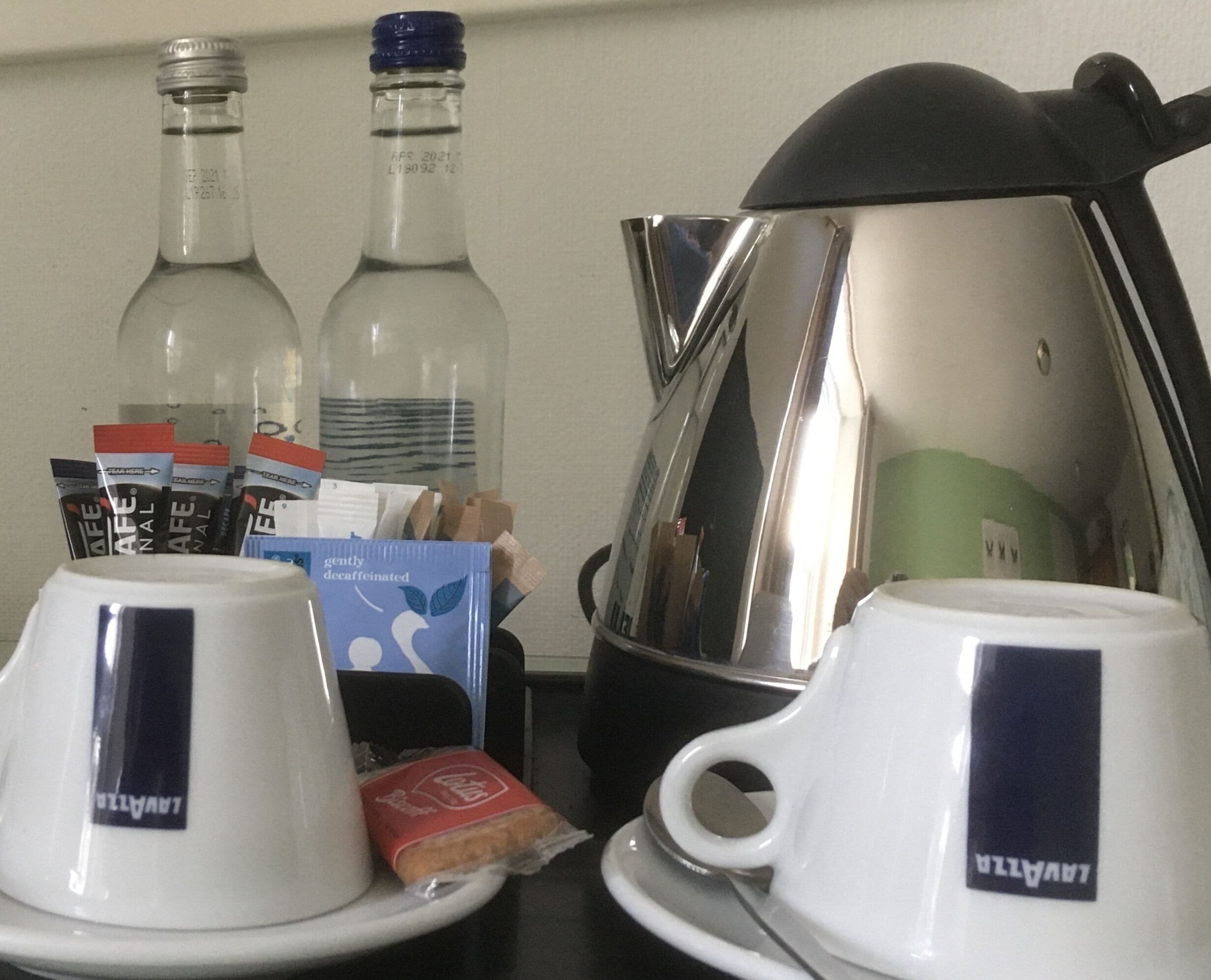 Una mesa con dos tazas blancas que dicen "LAVAZZA", una cafetera, dos botellas y algunos alimentos para el desayuno.