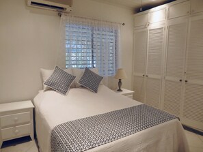 Un dormitorio con cama, dos almohadas, una mesita de noche con lámpara, un armario y una ventana con cortinas.