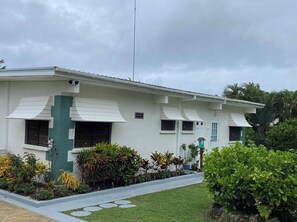 Edificio de dos pisos con techo plano, paredes exteriores blancas y detalles en verde. Se observan varias ventanas, una antena visible y un pequeño jardín con diversas plantas.