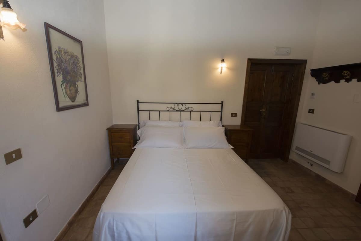 Quarto com cama, duas mesas de cabeceira, porta de madeira, radiador e quadro na parede.