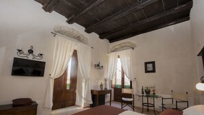 A bedroom with a large bed, wooden ceiling beams, two windows with white curtains, a small table with a vase, and a television on the wall.
