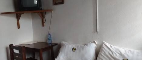 Quarto pequeno com uma cama, uma mesa de madeira, uma televisão sobre um suporte e uma garrafa de água na mesa.