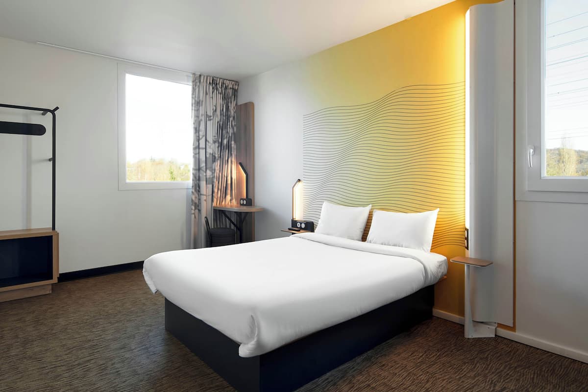 Una habitación de hotel moderna con una cama grande, una mesita de noche con lámpara, un armario y una ventana con cortinas.