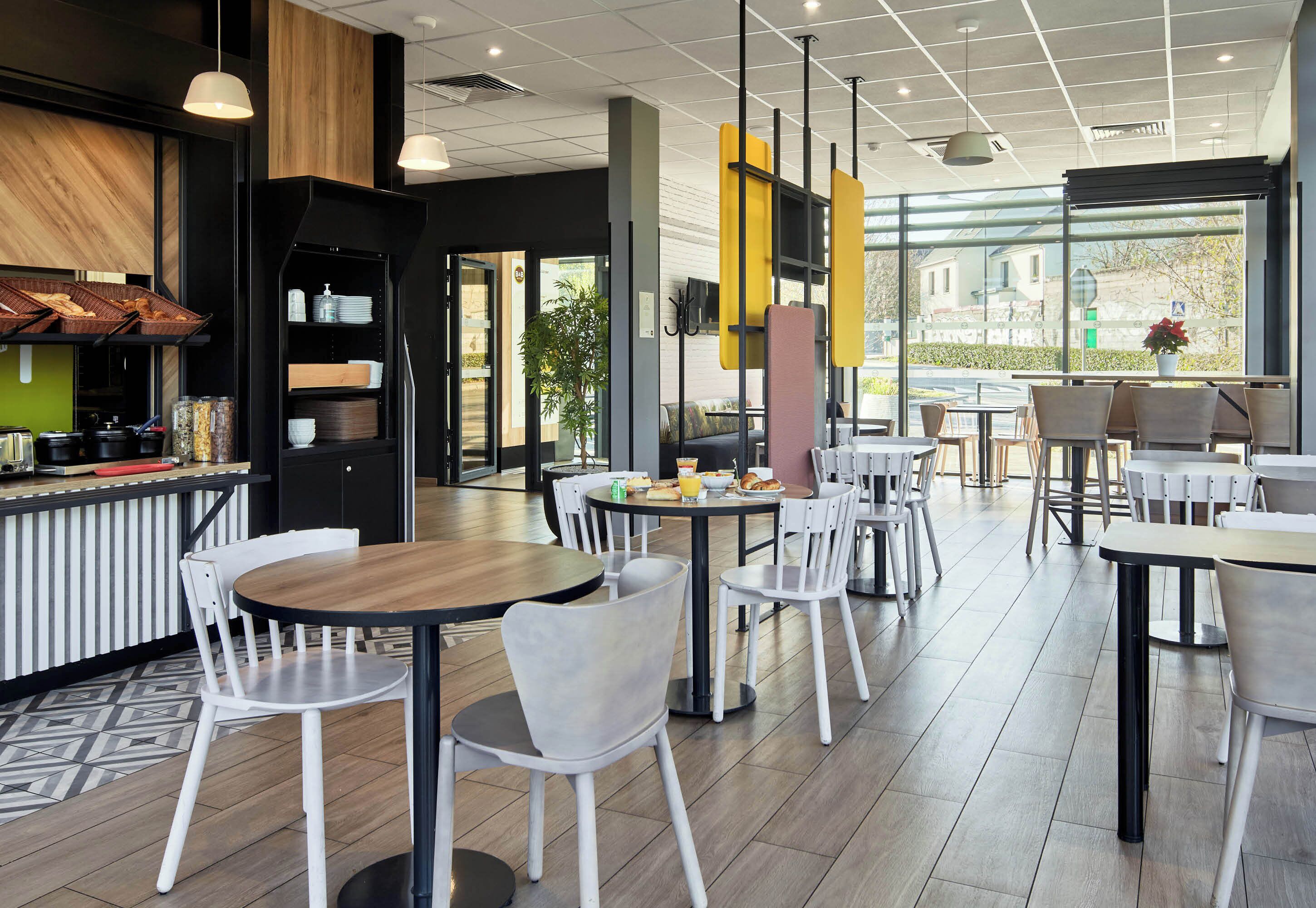 A modern cafe with round tables and white chairs, a kitchen area, and large windows offering a view of the outside.