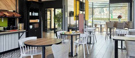 A modern cafe with round tables and white chairs, a kitchen area, and large windows offering a view of the outside.