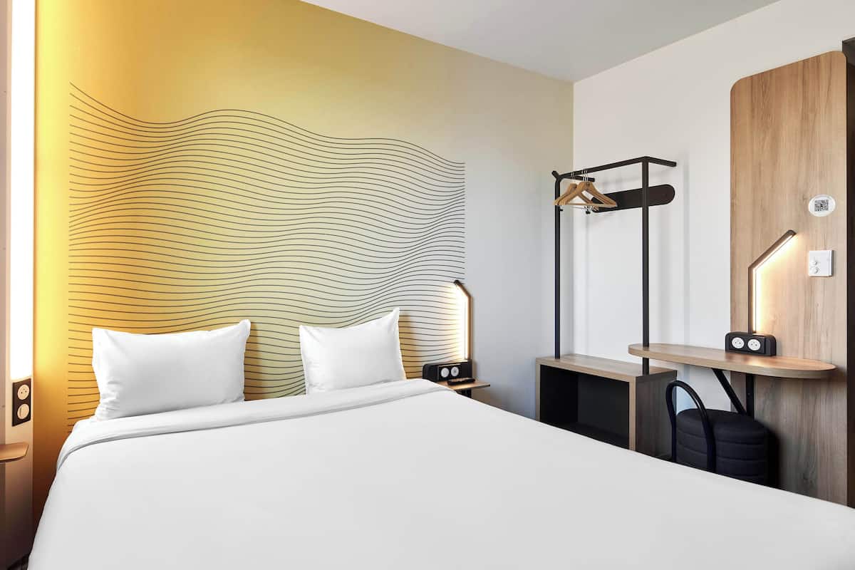 Una habitación de hotel moderna con una cama grande, un cabecero con un diseño ondulado, una mesita de noche con lámpara, un aparador de madera y un perchero.