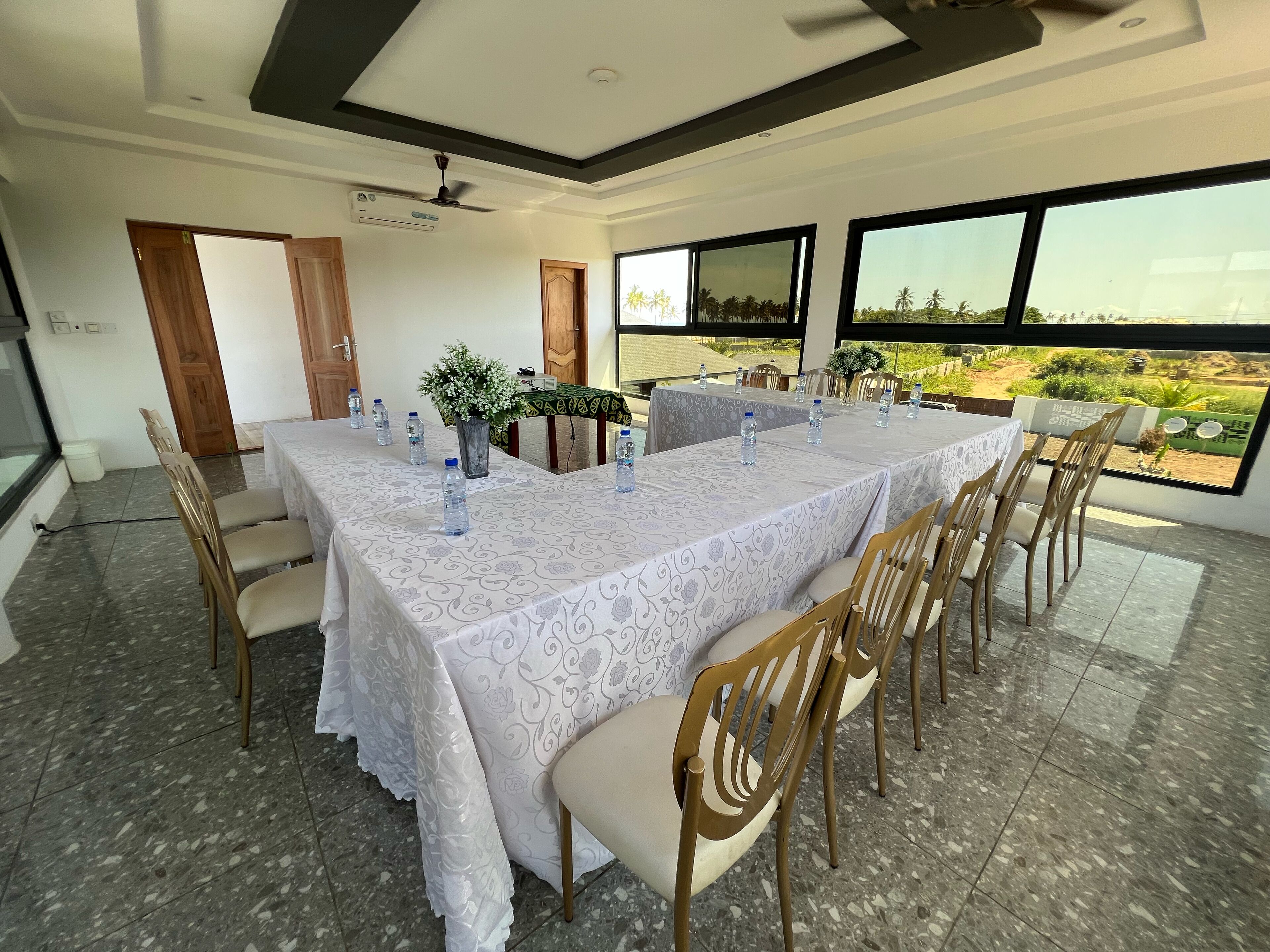 A dining area with a long table, chairs, and water bottles, with a view of greenery outside.