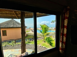 A room with a large window overlooking a pool, a thatched-roof building, and a palm tree.