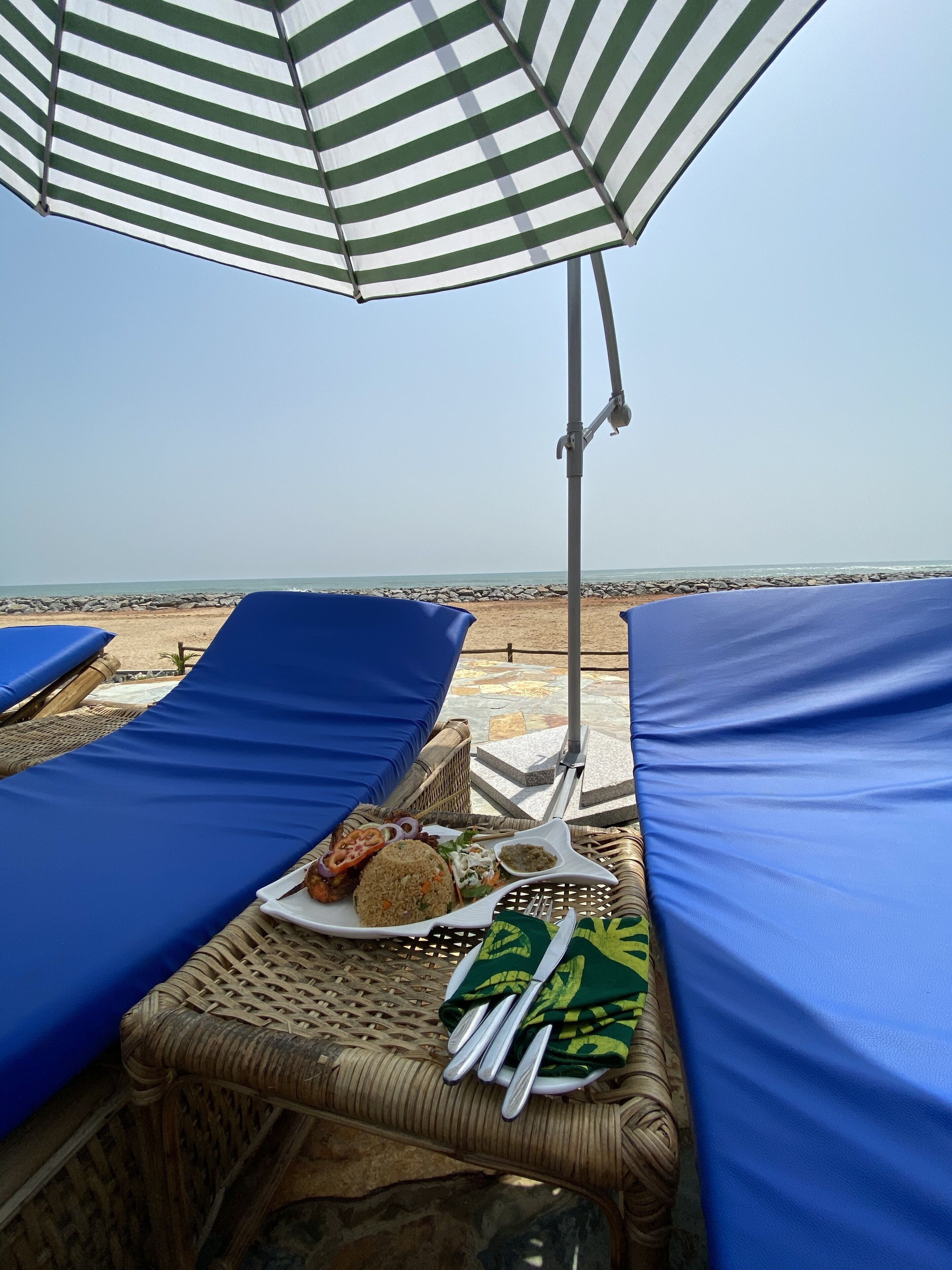 Un área de descanso junto a la playa con sillas acolchadas azules, una mesa con un plato de comida y una sombrilla a rayas.