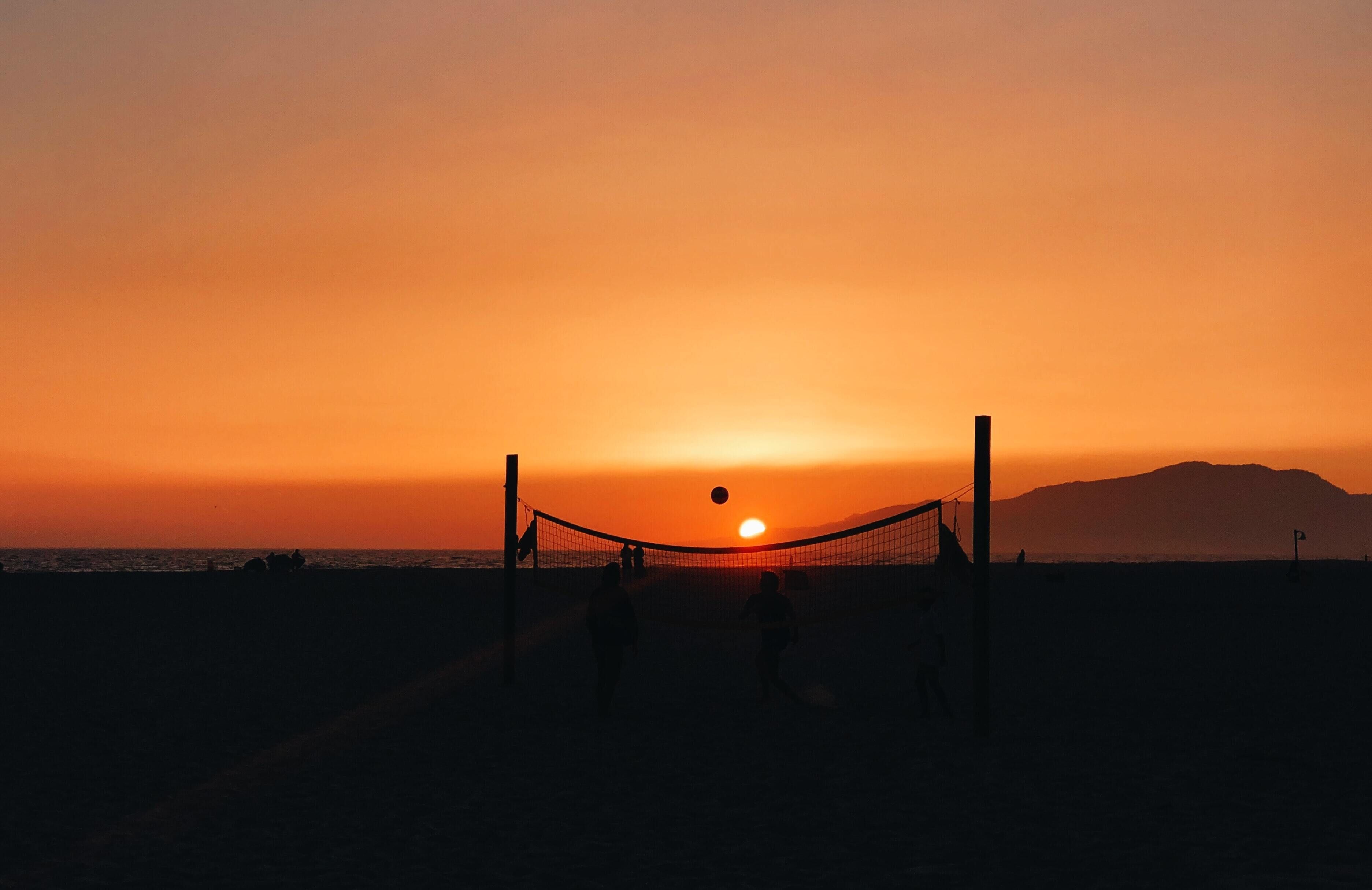 A beach volleyball net at sunset with silhouettes of people playing.