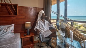 Habitación de hotel con un amplio ventanal con vista al océano, una cama con ropa de cama floral, una silla, una mesa con una copa de champán y una lámpara.