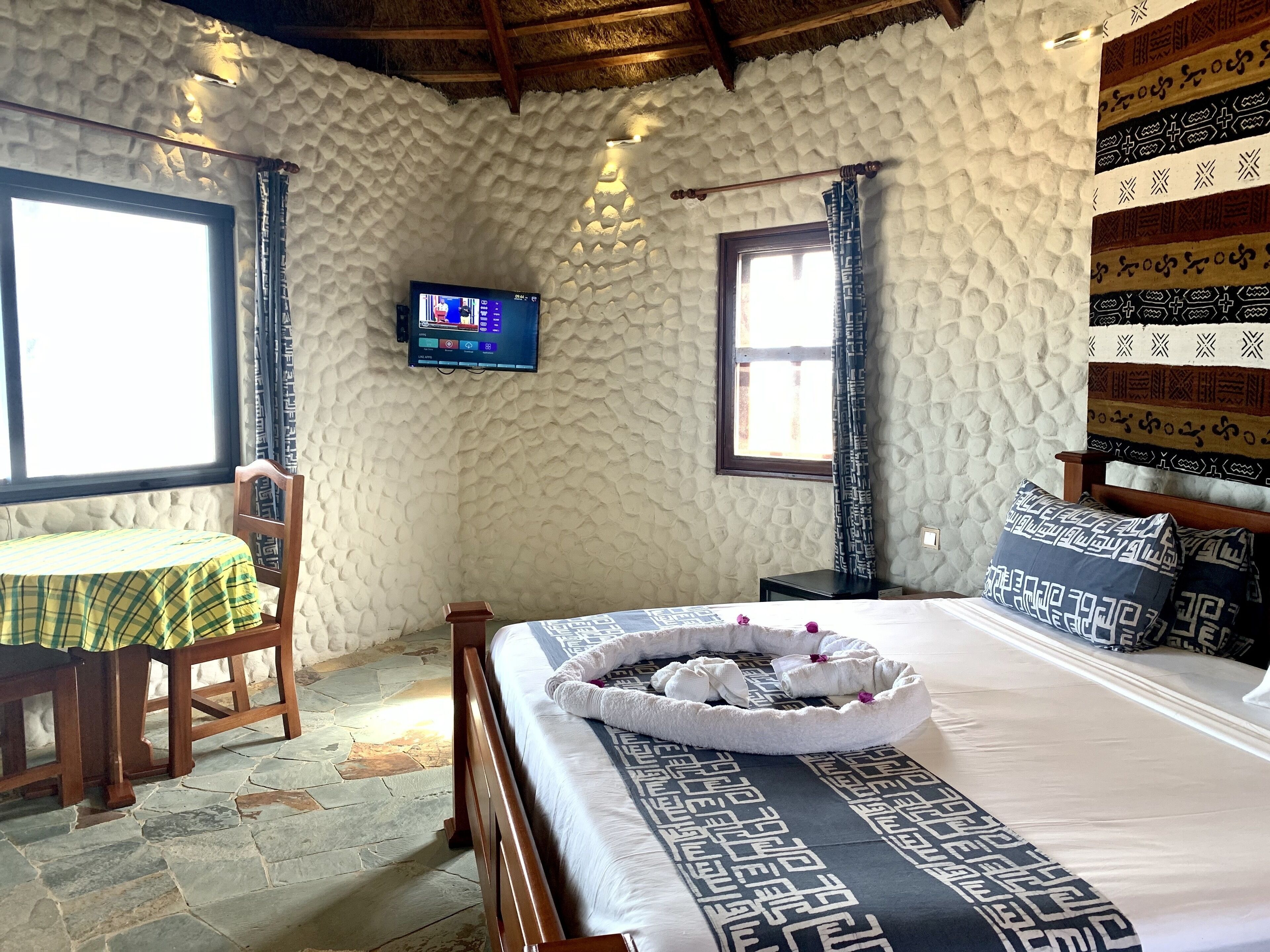 Een slaapkamer met een stenen muur, een bed met beddengoed met een patroon, een televisie aan de muur en een raam met gordijnen.