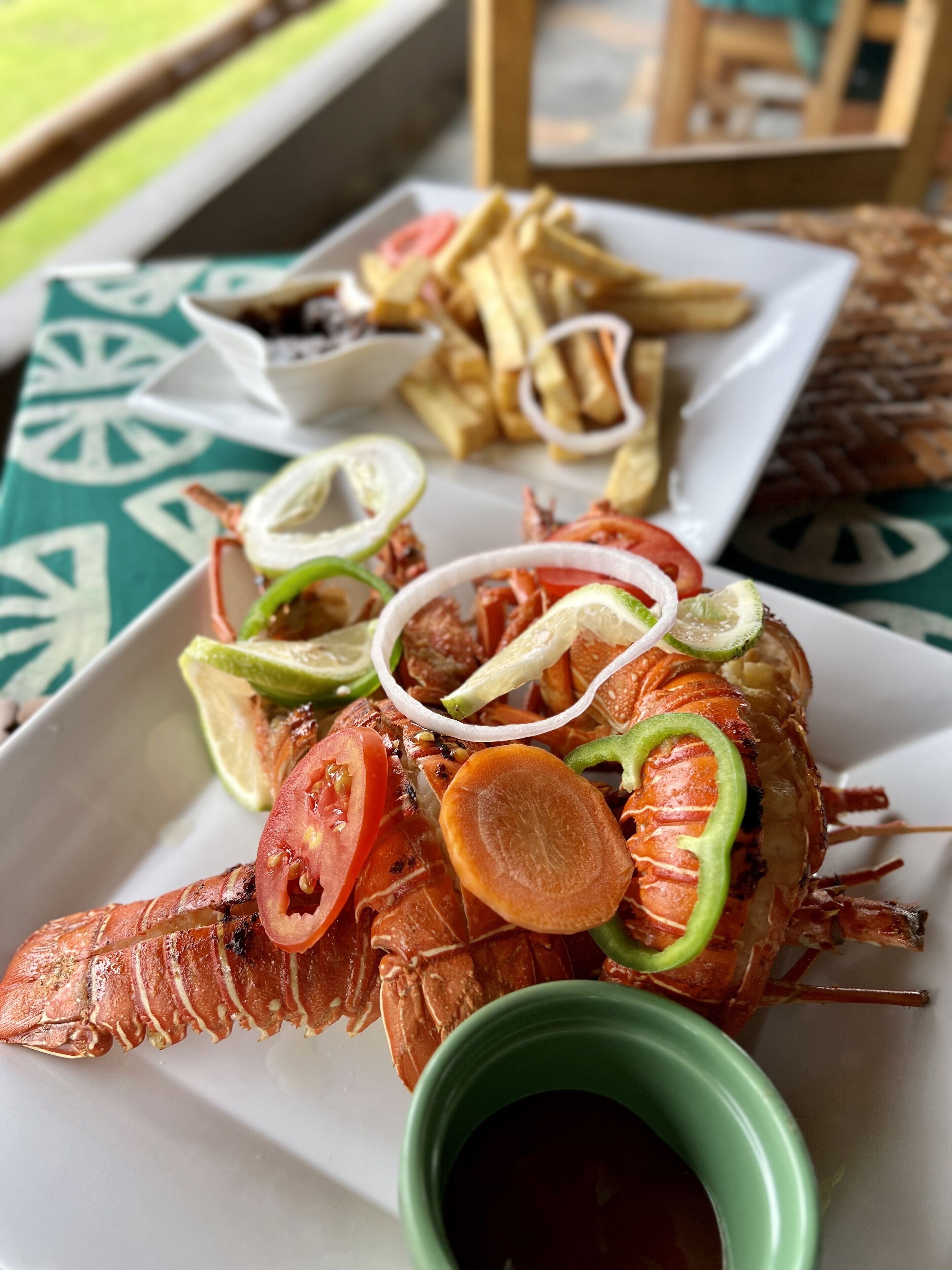 A plate of grilled lobster with lemon slices, tomatoes, and green peppers, accompanied by a small bowl of sauce and a side of fries.