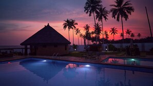 Una piscina con sillas de descanso y un edificio con techo de paja, rodeado de palmeras bajo un cielo al atardecer.