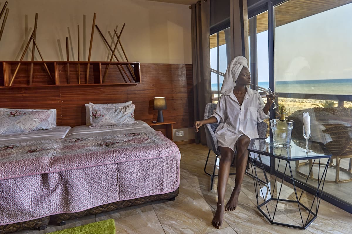 A bedroom with a large bed, a bedside table with a lamp, and a view of the beach through large windows.