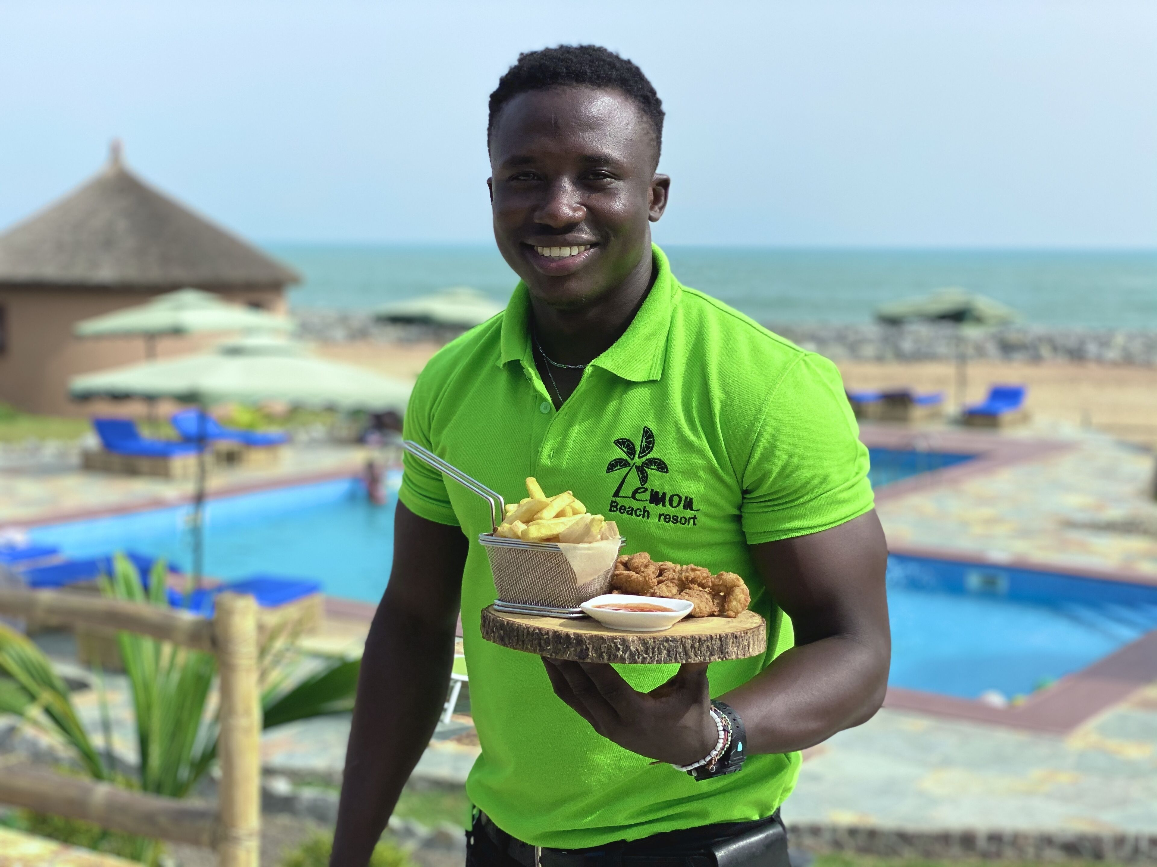 A person holding a tray with food and a drink, standing by a pool with lounge chairs and umbrellas.