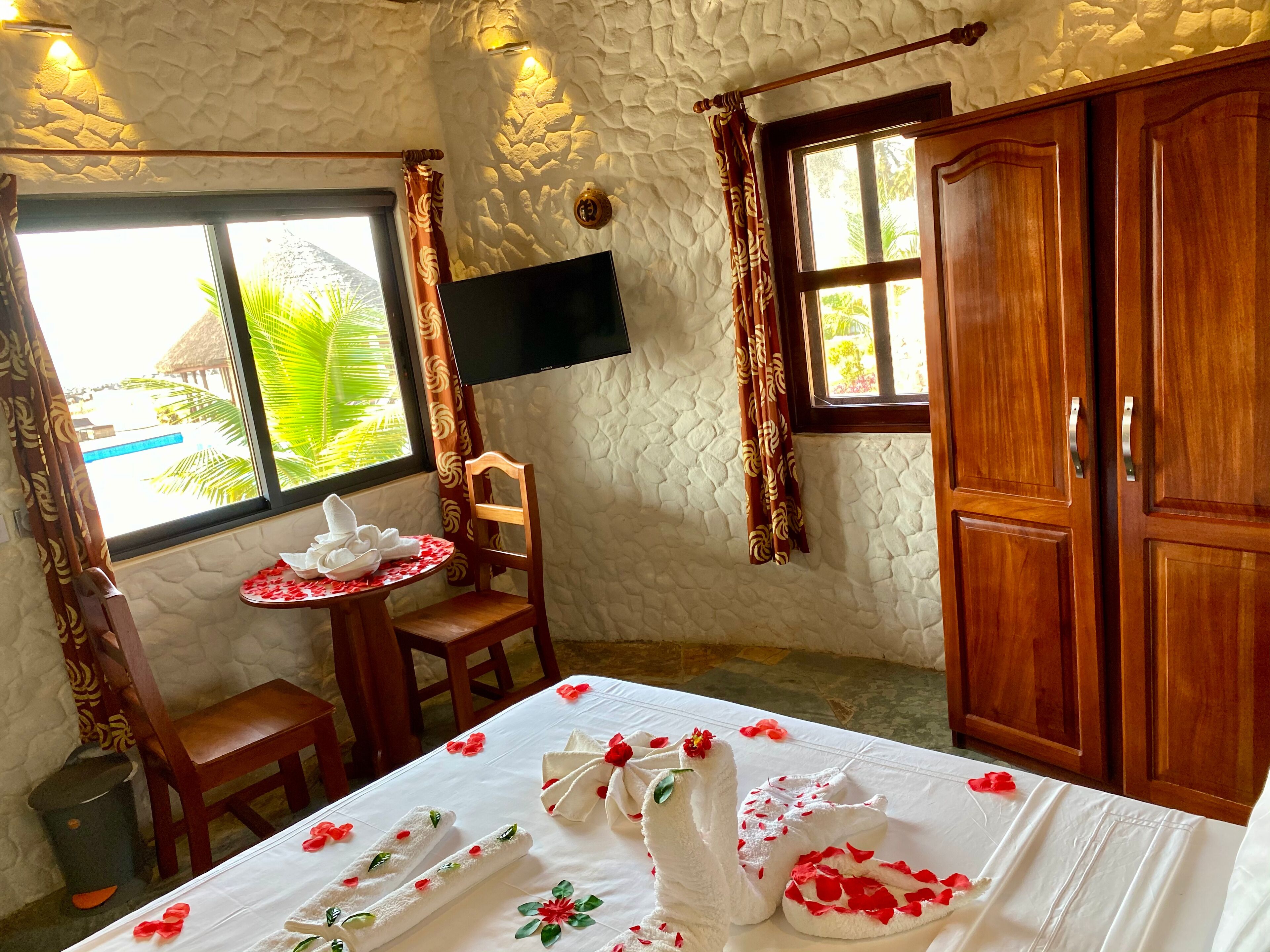 Una habitación con pared de textura de piedra, un mueble de madera, un televisor montado en la pared, una ventana con cortinas, una pequeña mesa redonda con una toalla y una flor, y una cama con una colcha blanca y toallas.