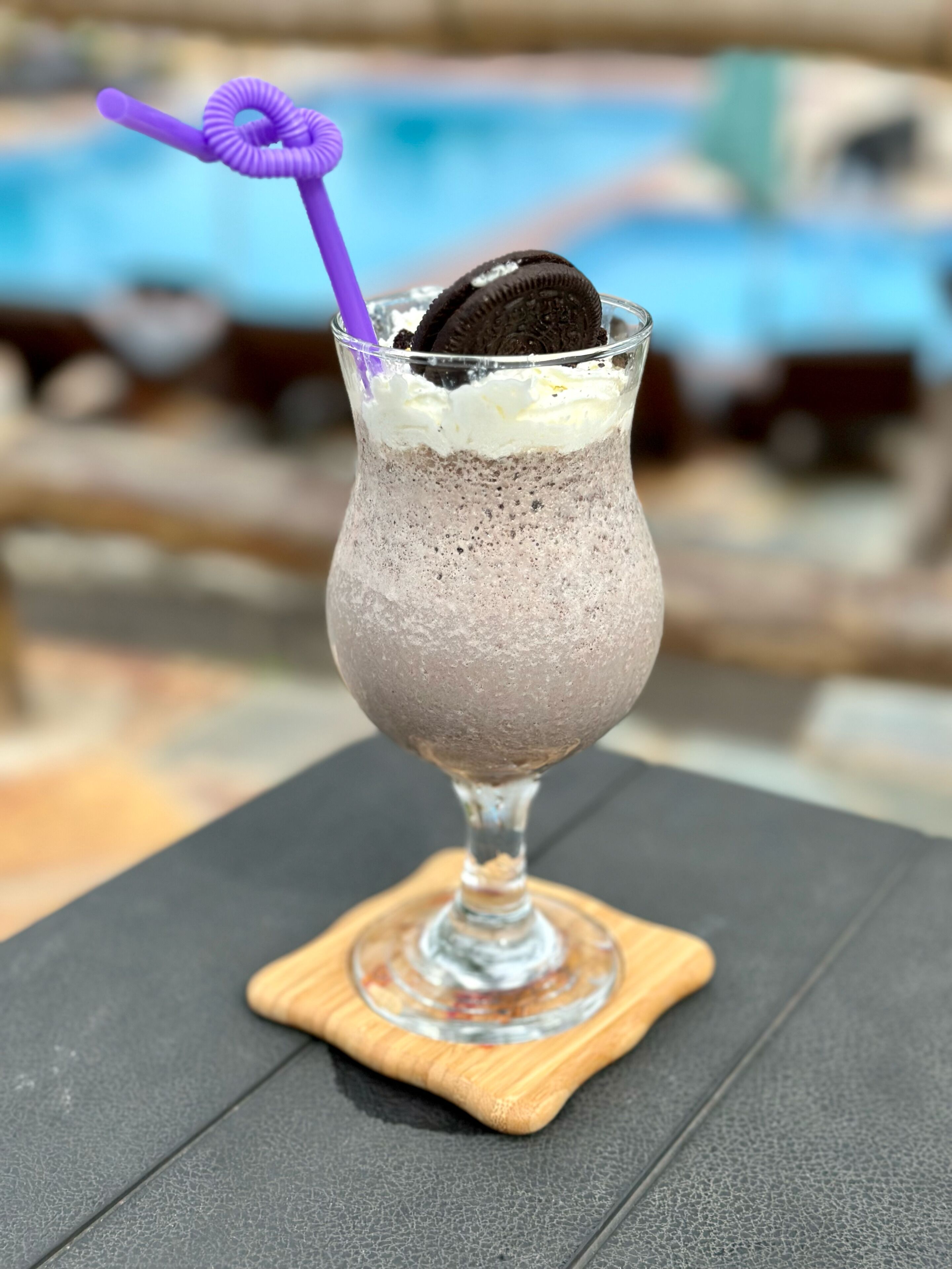 Un dessert servi dans un verre, accompagné d’une paille violette, le tout posé sur un sous-verre en bois.