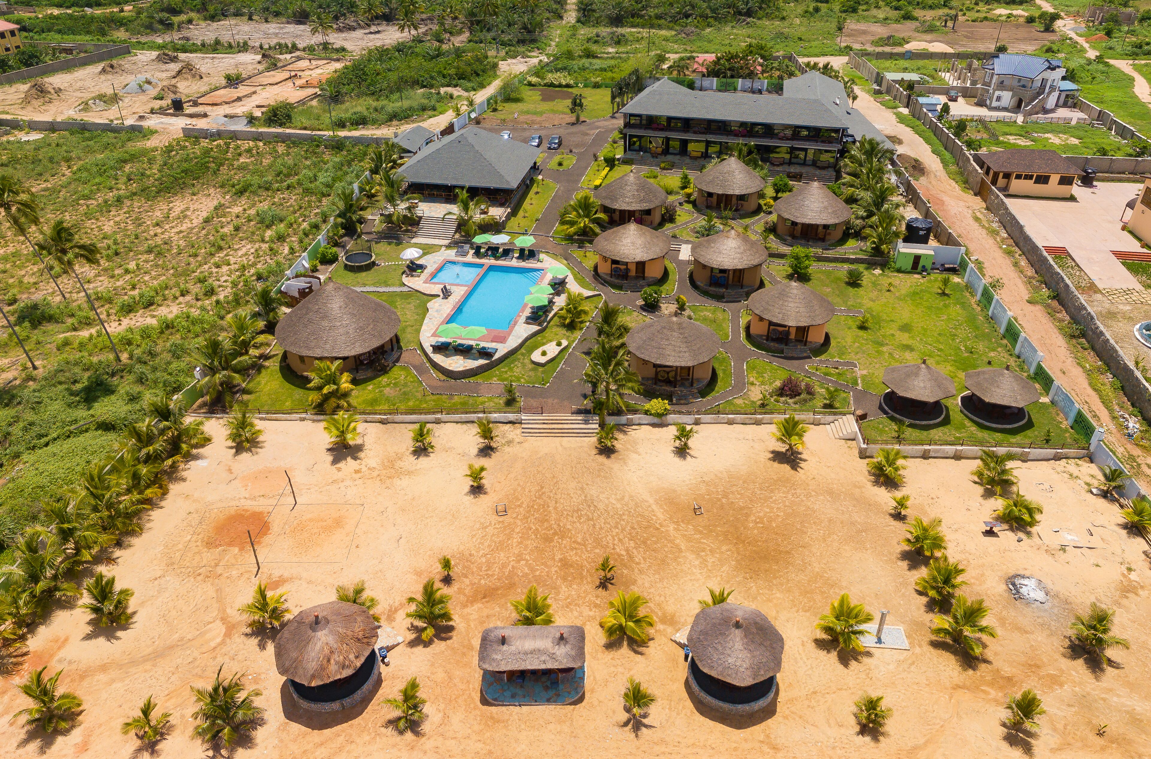 An aerial view of a resort with multiple thatched-roof huts, a swimming pool, and a large building likely serving as a main lodge.