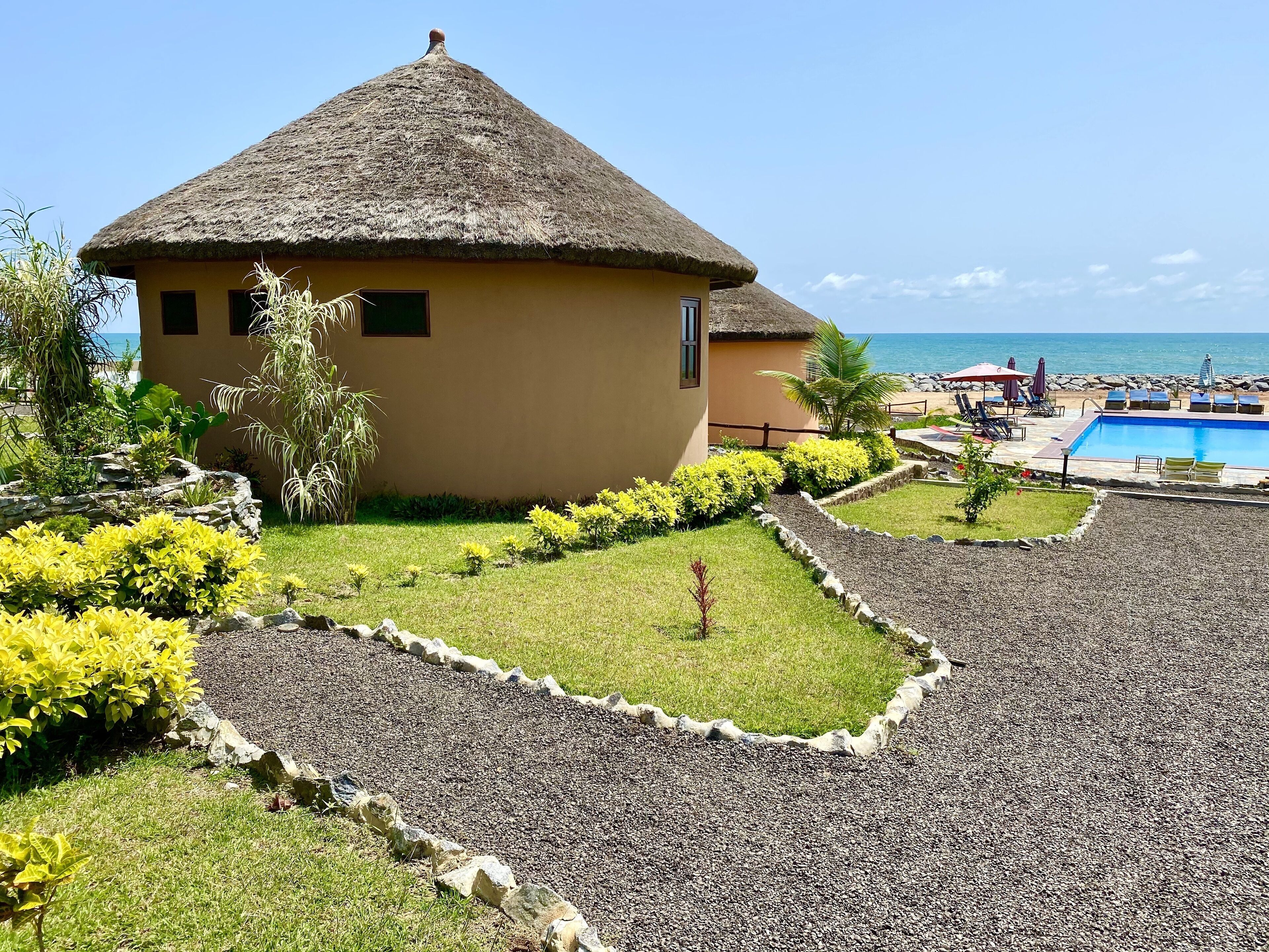 A thatched-roof building with a pool and lounge area, surrounded by well-maintained landscaping and a view of the sea.