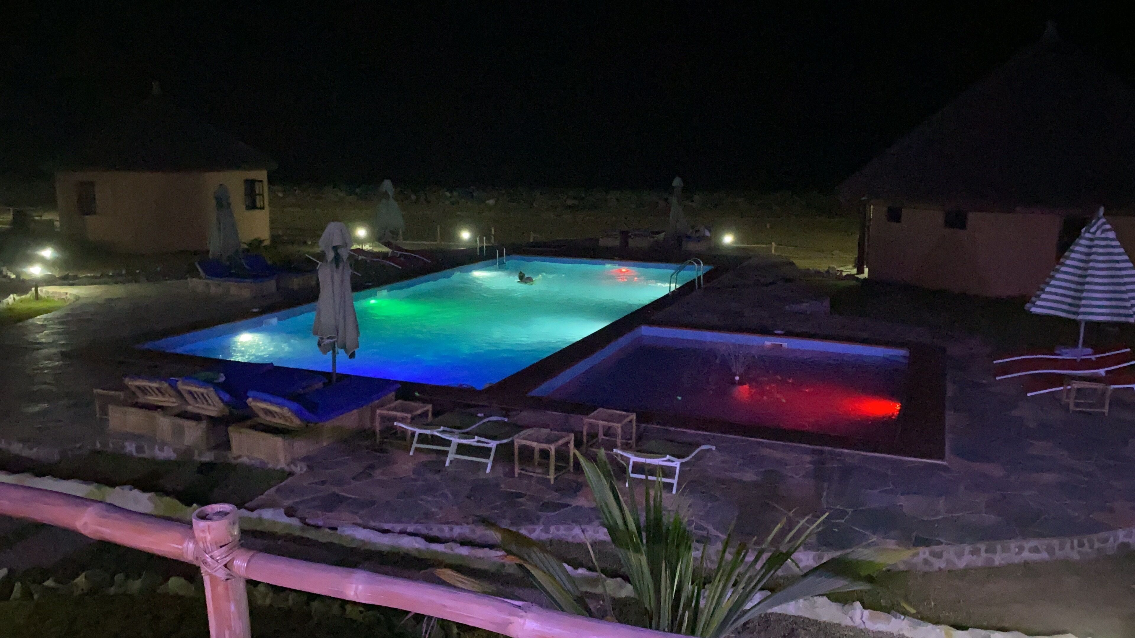 A swimming pool area with two pools, lounge chairs, and a small building.