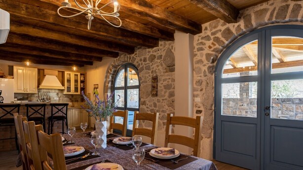 A dining area with a table set for four, wooden chairs, a vase of flowers, and a view of the outside through a stone wall and arched door. - Beautiful Villa Bogovski Dvori**** with a rustic touch on Krk (Malinska)