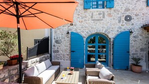 A stone building with blue shutters and a covered outdoor seating area with wicker furniture and an orange umbrella. - Beautiful Villa Bogovski Dvori**** with a rustic touch on Krk (Malinska)