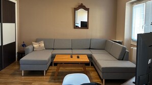 A living room with a grey sectional sofa, a wooden coffee table, a white chair, a mirror on the wall, and a television on the right.