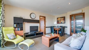 A modern living room with a sofa, a yellow armchair, a television, a bookshelf, and a dining area with chairs and a table. - Apartment Alcudia Portobello with seaview (Puerto de Alcudia)