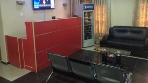 A reception area with a red counter, a television mounted on the wall, a black leather sofa, and a bar refrigerator. - Valton Hotels Abuja (Abuja)